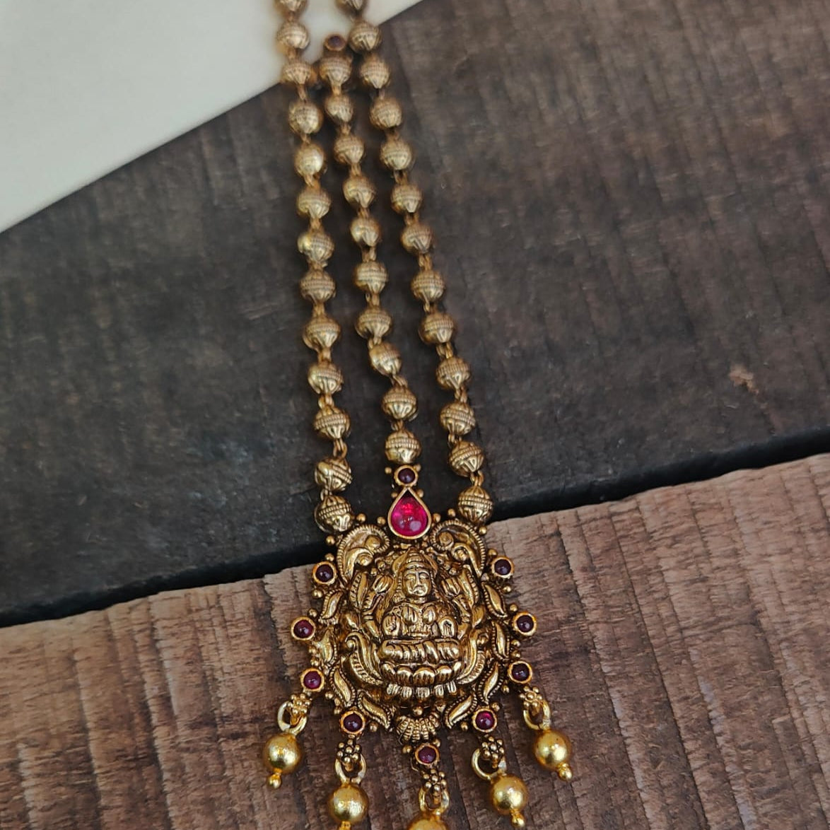 An antique-style gold-toned necklace featuring a detailed Ganesha pendant with a central red stone and dangling gold beads, displayed on a rustic dark wood background.